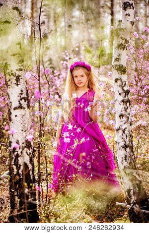 A Child With A Long Blond Hair Stands Among The Ledum And Birch In A Pink Dress And Smiling