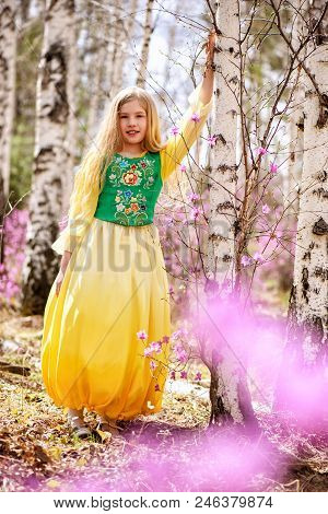 A Child With A Long Blond Hair Stands Among The Ledum And Birch In A Green Yellow Dress And Smiling