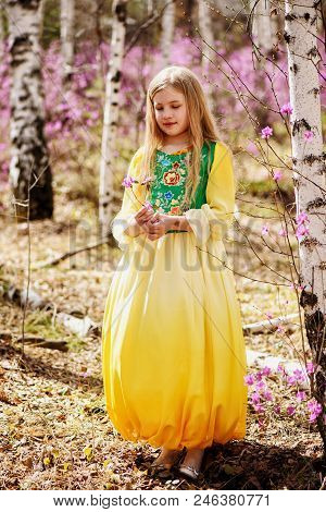 A Child With A Long Blond Hair Stands Among The Ledum And Birch In A Green Yellow Dress And Smiling