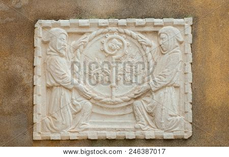 Religious Symbols In Venice. Crosier Staff Between Two Hooded Monks, An Ancient Stone Relief On A Wa