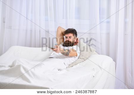 Man Laying On Bed, Watching Tv, White Curtains On Background. Guy On Serious Face Using Remote Contr