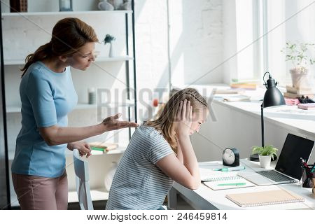 Depressed Teen Daughter Sitting At Work Desk At Home While Her Mother Yelling At Her