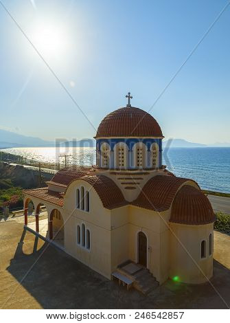 View Of Orthodox Greek Church At Mediterranean Coast. Orange Foof, White Building And Blue Sea Waves