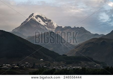 Georgian Village Stepantsminda On The Background Of The Snow-capped Mountain Kazbeg. Caucasus Mounta