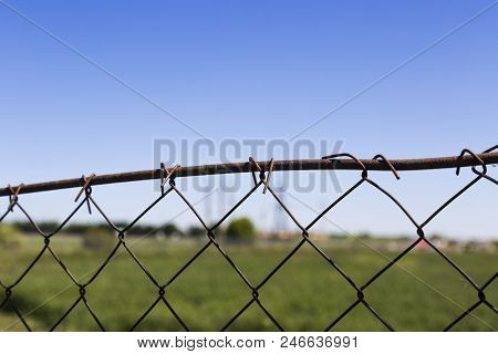 Old Rusty Wire Mesh Fence Against The Blurred Background Of A Green Field, An Grain Silos And A Clea