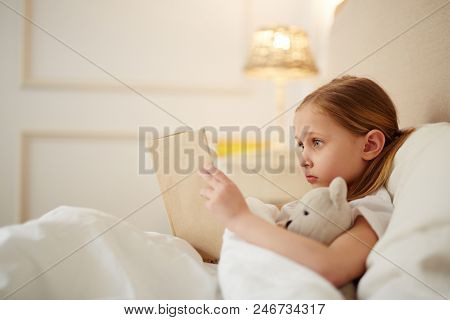 Cute little girl with teddybear lyiing in her bed and reading story tales before sleep