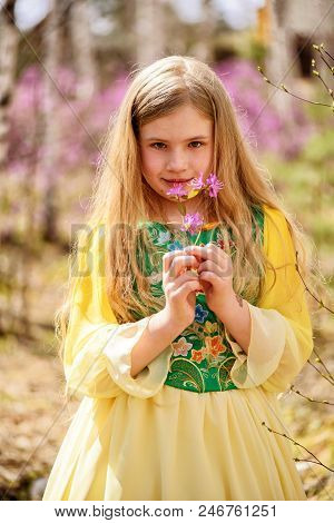 A Child With A Long Blond Hair Stands Among The Ledum And Birch In A Green Yellow Dress And Smiling