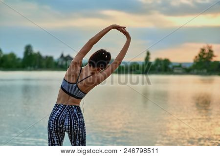 Backview Of Slim Girl Stretching Near Lake In The Evening. Sport, Yoga, Fitness. Model Maintaining H