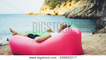 Young Hipster Relaxing On Coastline Beach On Inflatable Lazy Air Pouffe Sofa, Person Tourist Enjoy S