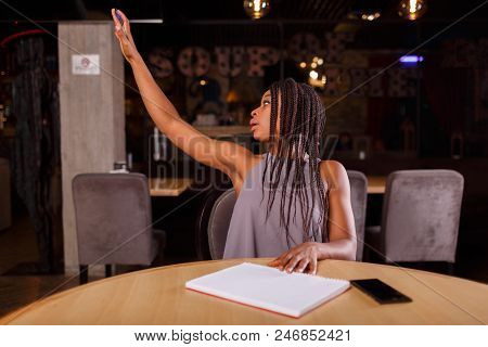 An Afro-american Woman Is Calling A Waiter With The Help Of Her Raised Hand.