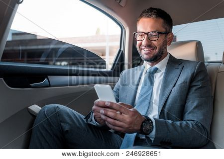 Smiling Businessman Using Smartphone On Backseat In Car