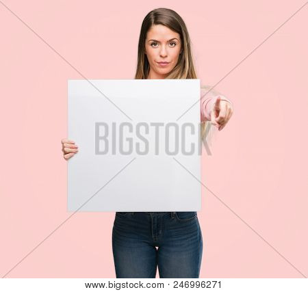 Beautiful young woman holding advertising banner pointing with finger to the camera and to you, hand sign, positive and confident gesture from the front