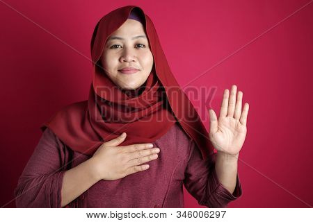 Portrait Of Happy Asian Muslim Woman Wearing Red Hijab Making Pledge Gesture, Hand On Chest Holding 