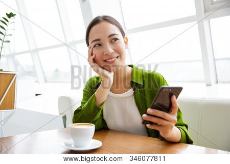Smiling asian woman using mobile phone while sittng at the cafe indoors and drinking coffee