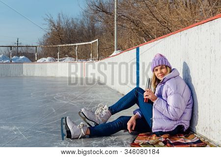 Lovely Young Woman Relaxing After Riding Ice Skates And Drinking Hot Drink From Termo Pot On The Ice