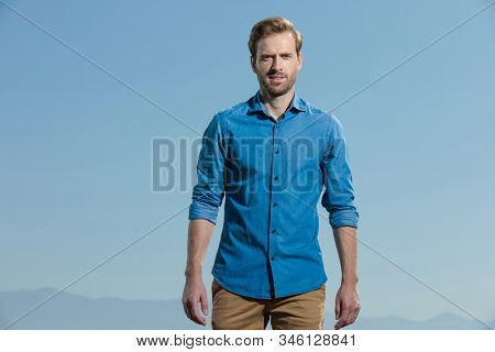 attractive casual man wearing blue shirt standing with hands loose and looking at camera happy outisde on blue sky