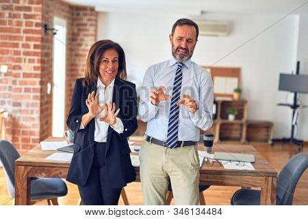Two middle age business workers standing working together in a meeting at the office disgusted expression, displeased and fearful doing disgust face because aversion reaction. With hands raised. Annoy