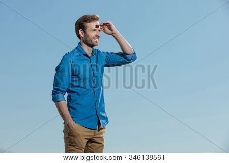 side view of a charming casual man wearing blue shirt standing with one hand in pocket and protecting his eyes with the other from the sun outisde on blue sky