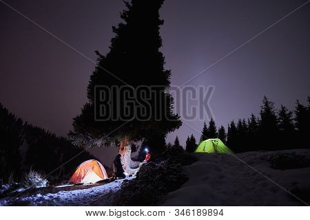 Man Sits Near A Tents At Night In The Winter In The Mountains
