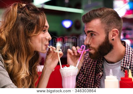 Photo of a cute positive young loving couple sit in retro bright cafe drinking cockail indoors.