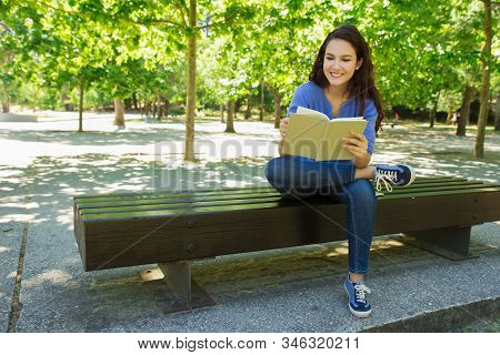 Beautiful Smiling Student Reading Book In Park. Attractive Young Woman Sitting On Wooden Bench In Pa