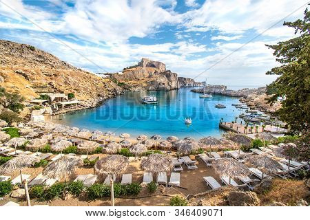 Lindos - View Of St. Paul Bay, Beach With Sun Beds And Umbrellas, Acropolis Of Lindos In Background 