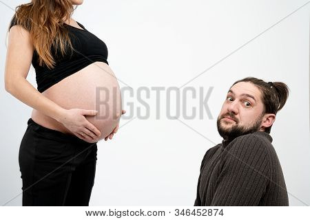 Embarrassed Young Bearded Man Looking At The Camera While His Pregnant Whife In Black Clothes Stands