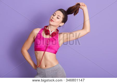 Horizontal Indoor Shot Of Joyful Well Kept Good Looking Fitness Trainer Holding Her Pony Tail In One