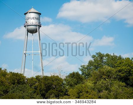Metal Water Tank Of Small Texas Town Gruene Standing High Above Trees On Sunny Day. Clouds And Blue 