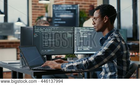 Portrait Of African American Developer Using Laptop To Write Code Sitting At Desk With Multiple Scre