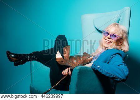 A Girl In A Blue Jacket And Purple Glasses Sits On A Chair. Fashion Photo In 80s 90s Style.