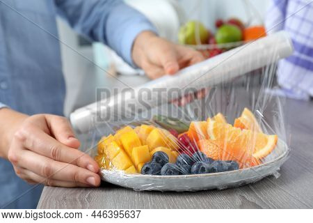 Woman Putting Plastic Food Wrap Over Plate Of Fresh Fruits And Berries At Wooden Table Indoors, Clos
