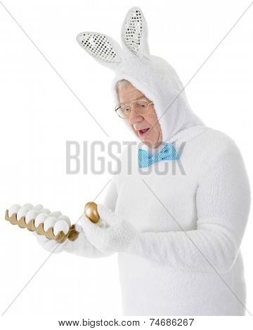 A senior adult looking surprised in his white Easter bunny outfit as he discovers a golden egg in the carton.  On a white background.