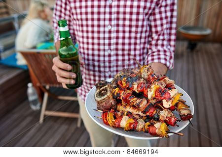 Man serves skewer kebabs for outdoor barbeque dinner party holding beer