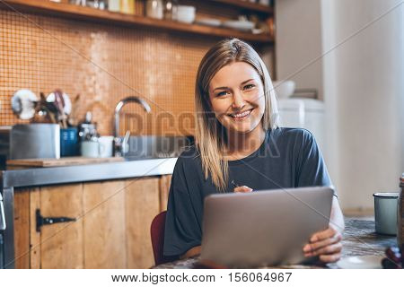 Portrait of an attractive young blonde woman sitting at a table in her kitchen using a digital tablet while eating breakfast