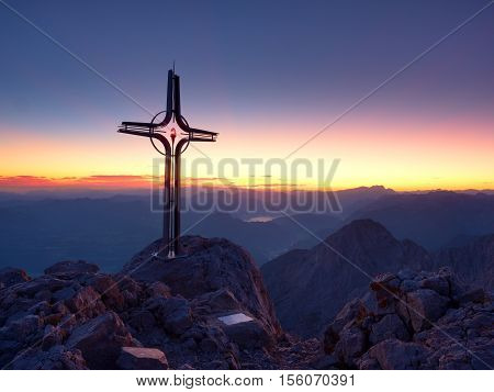 Steel Crucifix At Mountain Peak In Alps. Sharp Rocky Summit, Daybreak Sun In Sky.