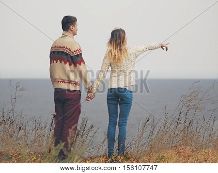 Young attractive couple in knitted sweaters standing on a cliff at sea shore and hugging girl pointing to a horizon with her hand