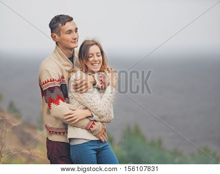 Young attractive couple in knitted sweaters standing on a cliff at sea shore and hugging