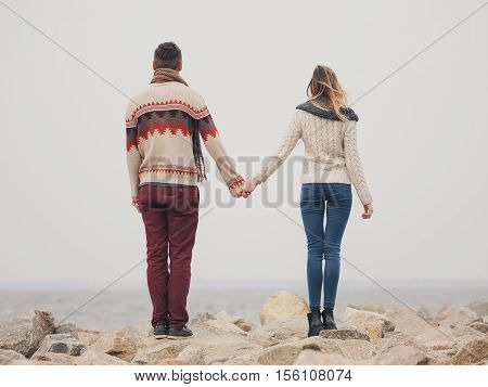 Young attractive couple in knitted sweaters on a cliff on sea shore hugging and holding hands creative toning