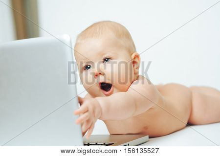 A baby with a surprised face lies in front of a silver laptop and holds out his hand