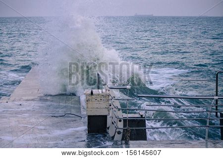 pier with Powerful sea splash wave with sprays and foam with vessel on the background. railings are broken because of huge wave.