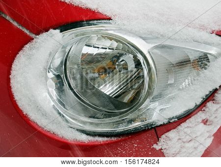 Detail of a red car with round headlights covered with snow during snowstorm