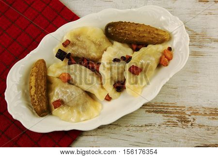 Boiled dumplings with meat topped with fried bacon with pickled cucumbers on white platter on an old wooden table. Traditional dish of Eastern Europe. Flat horizontal view from the top.