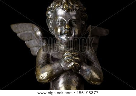 Praying winged putto on black background. Angel made of brass, covered with silver, as part of a candelabra from nineteenth century and symbol for religious passion. Macro object photo front view.