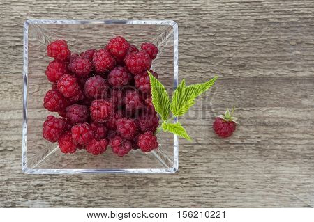 Organic raspberries in a glass bowl on a wooden background. Small green leaf.