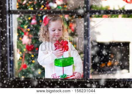 Family on Christmas morning at fireplace. Kids opening Xmas presents. Children under Christmas tree with gift boxes. Decorated living room with traditional fire place. Cozy warm winter day at home.