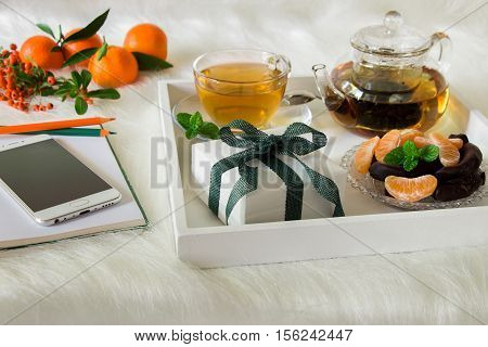 Green mint tea mandarin slices chocolate and gift box on tray near notebook pens mobile mandarins mountain ash sprig on white artificial fur background. Happy time break and gift. Close.