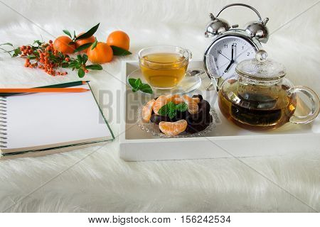 Green mint tea chocolate mandarin slices and alarm clock on tray near notebook pencils mandarins mountain ash on white artificial fur background. Morning time breakfast notebook plan preparation.