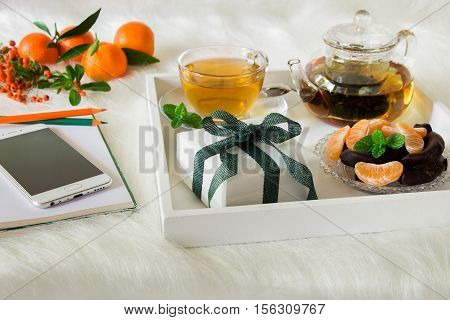 Green mint tea, mandarin slices, chocolate and gift box on tray, near notebook, pens, mobile, mandarins, mountain ash sprig on white artificial fur background. Happy time break and gift. Close.