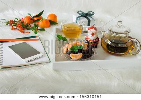 Green mint tea, chocolate, mandarin slices and Santa Claus on tray near mobile, notebook, pencil, mandarins, mountain ash on white artificial fur background. Time break Christmas surprise preparation.